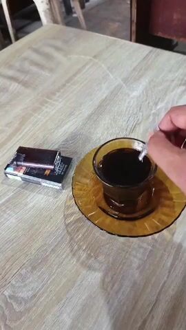 A top-down shot captures a pack of cigarettes and a lighter placed on a wooden table next to a glass of dark liquid, presumably coffee, with a person's hand stirring the liquid with a white object. The background is blurred but suggests an indoor setting.