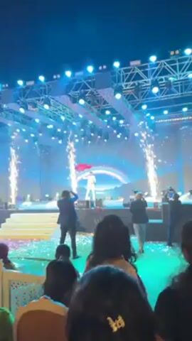 Here is a description of the video:
A wide angle shot shows a man dancing on stage at an event. He is wearing a white outfit with silver accents. Behind him, a digital backdrop displays a starry scene with a rainbow. Spark effects shoot up on either side of the stage. Stage lighting illuminates the scene in shades of blue and white. In front of the stage, a photographer in a blue suit takes pictures. Other people are visible in the foreground.