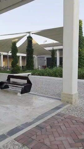 The video begins with a static shot of a paved walkway leading away from the camera. The walkway is bordered by gravel on the left and a brick pattern on the right. In the background, a white rectangular pillar stands prominently. To the left of the pillar, a large white canopy stretches overhead, supported by thin poles. Tall, dark green trees with conical shapes are visible beneath the canopy. Behind them, a building with windows and some white trim can be seen. A clock tower with a white face and black hands is visible in the upper right corner of the frame.

The camera then pans to the right, revealing a paved walkway where several people are walking away from the camera. A man in a blue and white tank top and black shorts is on the left, looking at his phone. A young girl in a floral outfit is walking slightly ahead of him. Behind them, other people are walking down the path, including a woman in a blue top. Palm trees and manicured green bushes line the path. A building with large windows and a tiled roof is visible on the right.

The camera continues to move, showing more of the palm trees, the green lawn, and the building. The building appears to be a restaurant or hotel, with "VENUS RESTAURANT" visible on a stone facade. Flowers, including purple and white blooms, are planted in beds along the building.

The video concludes with a shot looking towards the building and the garden area, with palm trees and manicured bushes creating a lush, tropical atmosphere. The sun appears to be setting, casting a warm light on the scene.