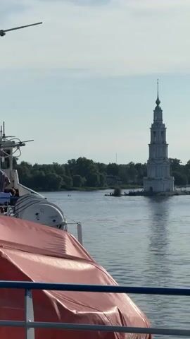 The video shows a tall, white bell tower partially submerged in water. The tower has multiple levels and a spire on top. There is water in the foreground, and the background shows trees and a cloudy sky. The camera zooms in on the bell tower. The view is from a boat, with a red and white cover visible in the foreground.