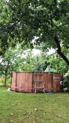 A wooden pool is shown in a backyard on a cloudy day. The pool is round with a metal ladder extending into it. The pool is surrounded by green grass and trees.
