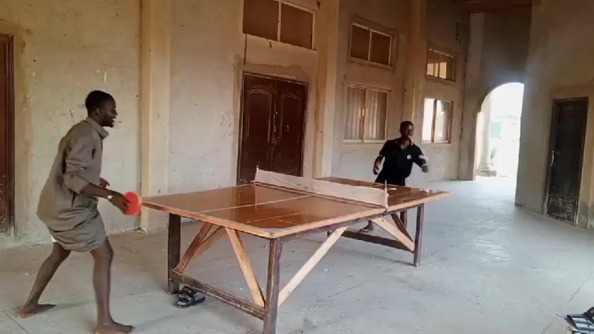 Two young men are playing table tennis in an indoor space with concrete walls and multiple windows. The table tennis table appears to be homemade, constructed with a wooden frame and a glossy, segmented playing surface. One man is positioned on the left side of the table, wearing a long-sleeved shirt and light-colored shorts, while the other is on the right, dressed in a black polo shirt. They are actively engaged in a game, striking a red ping pong ball back and forth. The environment seems somewhat aged and unfinished, with visible signs of wear and tear on the walls and the table itself. A pair of shoes are placed on the floor under the table. The setting gives the impression of a recreational space within a building that might be in a state of partial construction or renovation.