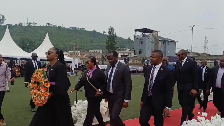 A group of formally dressed people walk on a red carpet laid over green turf, appearing to attend an event. The group is led by a woman carrying a large flower wreath. Most men in the group are wearing dark suits. The environment includes white tents, a building with a viewing platform, and a grassy hill in the background. Some individuals are looking directly at the camera, while others are engaged in conversation.