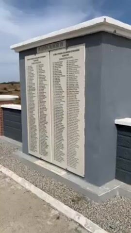 Here's a description of the video:

The video shows a memorial with two plaques listing names. The memorial is painted in shades of gray and white, with a brick base. The plaques are white with black lettering. The text at the top reads "In Loving Memory Of". Behind the memorial is a landscape with low, dry hills under a blue sky with some clouds.