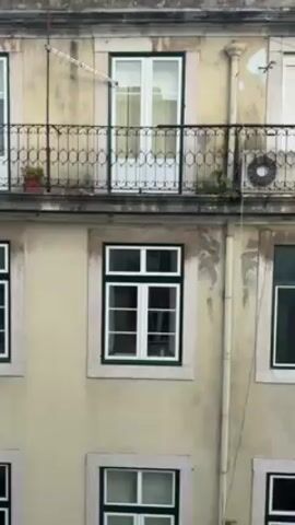 The video shows the facade of a multi-story building. The building has several windows with white frames and green or black trim, some with balconies. The facade is painted in different colors, including light yellow and white. The building's roof is red-tiled. The sky is visible in the background, with some clouds. The video zooms out slightly to reveal more of the building and neighboring structures.