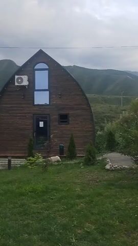 The video begins with a close-up shot of a wooden house with a dark-colored roof. An air conditioning unit is mounted on the left side of the house. There's a large arched window and a smaller rectangular window on the front of the house. A dog is lying on the grass in front of the house, and there are some small trees planted in the yard. The camera then pans to the right, revealing a hilly, green landscape with trees and some mountainous terrain in the background. The sky is overcast with clouds. In the distance, on top of a hill, a person can be seen standing. The video appears to be shot outdoors in a natural setting.