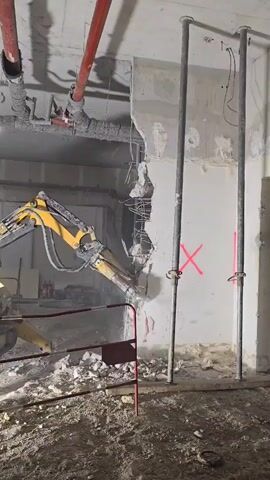 The video shows a demolition scene. A yellow hydraulic excavator with a jackhammer attachment is breaking down a concrete wall. The wall is reinforced with rebar, visible in the broken section. Two vertical metal support beams are in front of the intact portion of the wall, marked with red X's. Red pipes run along the ceiling above. The floor is covered in rubble and dust from the demolition. A red metal barrier is positioned to separate the work area from the viewing area.