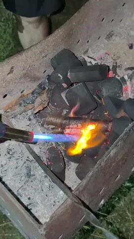 This is a close-up video of someone using a blue and orange flame torch to ignite charcoal in a metal grill. The charcoal is piled in the grill, and some pieces are already glowing red and emitting flames. The background is blurred, but appears to be an outdoor setting with grass. A person's legs and feet, wearing dark pants and tan boots, are partially visible at the top of the frame.