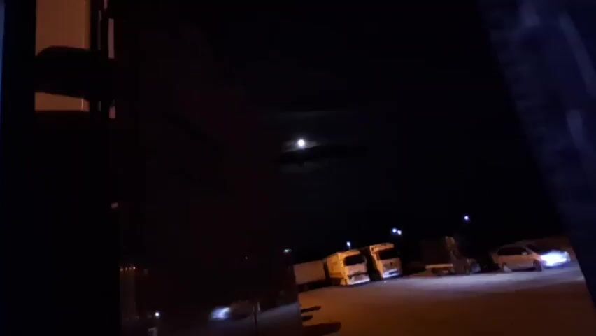 The video begins by showing a nighttime scene, likely a parking lot or industrial area, with several parked trucks and cars illuminated by artificial lights. The moon is visible in the dark sky, partially obscured by clouds. A large building with text on its side is in the foreground on the left. The camera then zooms in on the moon, which appears bright and is surrounded by drifting clouds, creating a somewhat atmospheric view. There are no people visible in the video.
