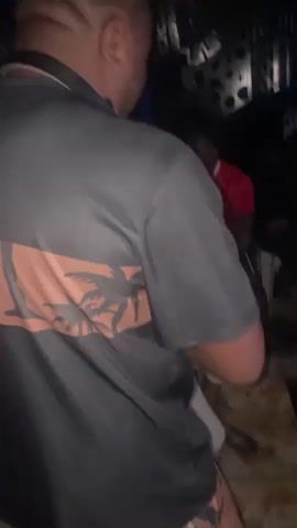 The video shows a group of men in a dimly lit room, possibly a club or lounge. The camera is focused on the back of a man wearing a gray t-shirt and a black collar. He is looking down and appears to be holding something in his hands. In the background, other men are visible, some sitting and holding drinks. The room has decorative elements on the walls, which are illuminated by blue lights. The overall atmosphere seems to be casual and social.
