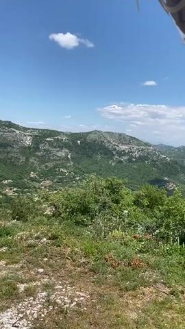 This is a first-person perspective video recorded on a sunny day in Lipa Dobrska, Montenegro. The scene is a lush, mountainous landscape with green trees and vegetation covering the hills. The sky is a clear blue with a few scattered white clouds.

The camera is positioned inside what appears to be a vehicle, possibly a small tour bus or tram, as evidenced by the clear plastic canopy and a white pole extending into the frame. A hand, wearing a purple bracelet with white text that reads "LARA", is visible on the right side of the frame, holding onto a green object. The vehicle is moving, as indicated by the slight blur of the surrounding greenery and the shifting perspective.

The activity appears to be a scenic tour or journey through the Montenegrin countryside. The overall mood is peaceful and relaxed, capturing the beauty of nature on a bright, clear day. The mountainous terrain suggests a rural or remote location, characteristic of many parts of Montenegro.