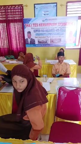 The video shows a classroom setting during a "TKA (TES KEMAMPUAN AKADEMIK)" or "Academic Ability Test," as stated on a banner at the front of the room. The banner also indicates that the test is for elementary school students of class 6, and the date is July 14. Several students are seated at individual tables, facing forward. Some students wear school uniforms, while others are in casual attire. The tables are covered with yellow tablecloths. A woman wearing a brown hijab is partly in the foreground, with her back mostly to the camera. The room is brightly lit and has red and maroon curtains on the windows. There is a framed poster or document on the wall above the banner.