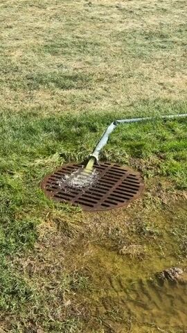 The video shows a close-up, overhead shot of a large, rusty metal grate on the ground, partially submerged in murky, yellowish water. Green grass surrounds the grate, with some patches of dry, brown grass also visible. A grey hose, reinforced with a white sleeve, is bent and inserted into the grate, with a stream of clear water gushing out of it. The water flows over the grate and into the puddle below. The lighting suggests it is daytime, with the sun casting shadows. The scene appears to be an outdoor environment, possibly a field or park, where water is being drained or pumped. There are no people visible in the video.