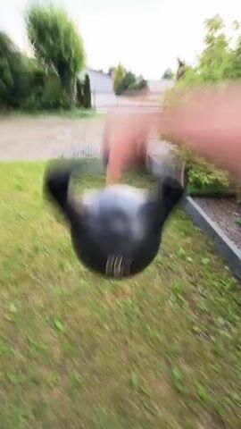 The video shows a person's hand reaching for a black kettlebell. The kettlebell has "20 KG" visible on it. The scene is outdoors, with green grass and some foliage in the background. The camera is in motion, giving a shaky, first-person perspective. The person is wearing dark pants and bright blue socks.
