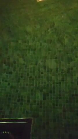 A man stands in a green-lit pool within a cave-like structure.  Water pours from above, creating many ripples and bubbles on the surface of the pool. The pool’s bottom is covered in a green tile mosaic. Light reflects from the water, illuminating the cave’s ceiling and walls, creating a dark atmosphere. The man is partially submerged, facing away from the camera.