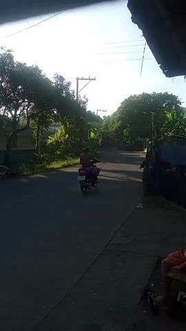 The video shows a person riding a motorcycle down a street. The sun is shining brightly, casting shadows on the road. There are trees and houses on either side of the street, and power lines are visible overhead. The person on the motorcycle appears to be wearing a helmet and a backpack. The video is shaky and the quality is not very good.