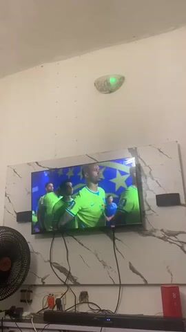 The video shows a television screen displaying a group of men in green and yellow uniforms standing in front of a flag with yellow stars on a blue background. The men appear to be soccer players. They are looking to their right, with their heads slightly bowed. The television is mounted on a wall with a marble-patterned backdrop. Below the television, there is a black soundbar, various cables, an electrical outlet, and a fan. The room appears to be dimly lit.