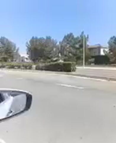 The video shows a view from inside a vehicle. The vehicle appears to be moving along a paved road. On the right side of the road, there is a line of trees and bushes, with some buildings visible in the distance behind them. The sky is clear and blue. In the foreground, the side mirror of the vehicle is visible on the left side of the frame. There is no visible activity of people or any interaction. The scene appears to be a suburban or commercial area.