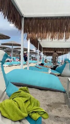 The media file depicts a sunny day at a beach resort in Paralia Mesis, Greece. The scene is set under a canopy with thatched roofing, providing shade to a seating area with large, turquoise beanbag chairs and low tables. A black backpack rests on one of the tables. In the foreground, a lime green towel is draped over a turquoise and white beanbag chair.

Beyond the shaded area, the beach is lined with rows of identical turquoise sun loungers and parasols with natural straw canopies, characteristic of a Mediterranean beach. Several people are visible, some reclining on the loungers, while others are standing or walking. One woman in a dark dress walks between the loungers, and a man in a tan uniform stands near the back of the seating area. Two seagulls are present on the sand, one near a lounger and another further back.

The sky is blue with scattered white clouds, indicating pleasant weather suitable for a beach outing. The sea is visible in the background, calm and stretching towards the horizon. The overall atmosphere is relaxed and leisurely, suggesting a vacation or a day of leisure. The presence of numbered signs on the beach, such as "A10" and "A19", indicates a structured resort or beach club environment. The time of day appears to be daytime, likely late morning or early afternoon, given the bright sunlight and shadows.