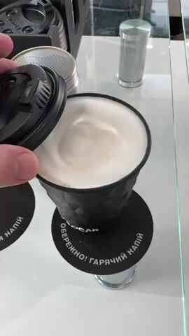 A hand places a black lid on a black paper coffee cup. The cup has a textured diamond pattern. A black circular coaster with white text reads "ОБЕРЕЖНО! ГАРЯЧИЙ НАПІЙ" in Cyrillic script, which translates to "CAUTION! HOT BEVERAGE." The cup sits on a white surface next to a glass table. In the background, there are other coffee lids and a silver cylindrical object.