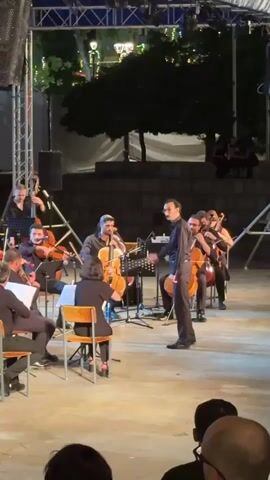 A video shows an outdoor orchestra performance at night. The orchestra is composed of about 10 musicians playing string instruments, including cellos and violins. The conductor, wearing a dark suit, stands in front of the orchestra and gestures with his hands. The musicians are seated, and the conductor is standing. They are performing on a stage with a backdrop of trees and lights. An audience is visible in the foreground, with their backs to the camera. The overall atmosphere appears to be a formal concert.