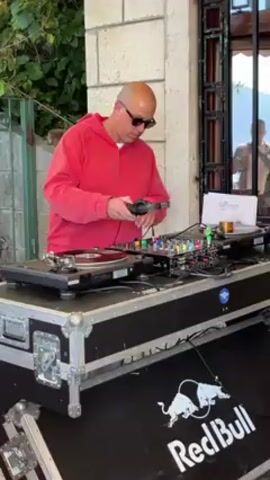 A male DJ stands behind a DJ setup, wearing a red sweatshirt and sunglasses. He’s bald and focused on adjusting the settings of his equipment. He’s operating two turntables and a mixing board, which are set on a large road case bearing the Red Bull logo. The location appears to be outdoors, possibly on a patio, with a stone wall and foliage visible in the background.  A reflective glass door is behind the DJ.