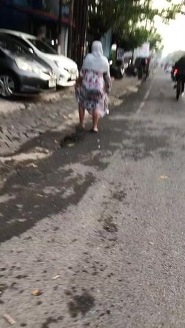 A person wearing a light-colored hijab and a floral dress walks away from the camera on a wet, asphalt road. The person is wearing dark-colored shoes and appears to be middle-aged. The person is walking on the right side of the road. To the left of the road, there are two cars parked on a cobblestone surface. The cars are dark gray and white. In the background, there is a building with blue and yellow elements, and some trees. The lighting suggests it is daytime, possibly late afternoon or early morning due to the soft light. There are some visible water puddles and wet patches on the road, indicating recent rain or street cleaning. The overall scene appears to be an urban or semi-urban environment.