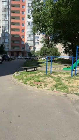 The video pans across a residential courtyard on a sunny day. In the foreground, a paved driveway curves to the left, bordered by a grassy area with a wooden bench and a children's playground structure with a blue frame and a green slide. Across the driveway, several cars are parked in a row along the side of a tall, multi-story apartment building. This building has a light gray facade with sections of orange and balconies. To the left of this main building, another similar apartment building is partially visible, with the same architectural style. Further in the background, more apartment buildings and trees can be seen. The sky is clear and blue.