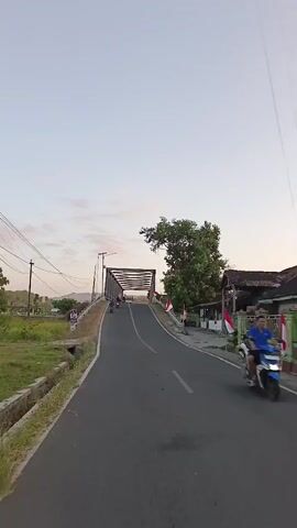 The video shows a road leading to a bridge. The road is paved and has white lines marking the lanes. There is a grassy area with a ditch on the left side of the road. On the right side of the road, there are some buildings and trees. Two Indonesian flags are visible on poles near the bridge. There are a few people on motorcycles on the bridge, and one person is walking. The sky is a light blue color, and the sun is setting. The overall scene is peaceful and rural.
