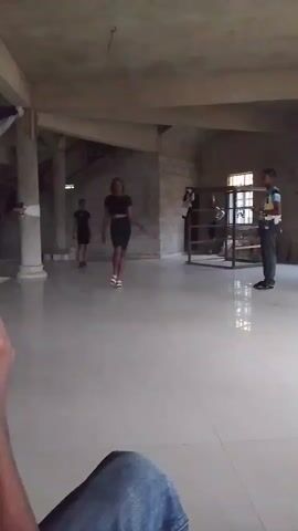 In an interior space featuring gray tiled floors and concrete ceilings supported by pillars, a woman in a black crop top and knee-length black skirt walks directly towards the camera, striking a pose with her hands on her hips. To the left of the frame, a young boy in a black outfit stands, while to the right, a person in uniform looks on. Two more figures stand near a wooden barrier behind them. A person's arm, clad in a blue denim sleeve, is visible at the bottom left of the frame. As the woman poses, the camera moves slightly, and another arm holding a smartphone appears, presumably capturing the scene.