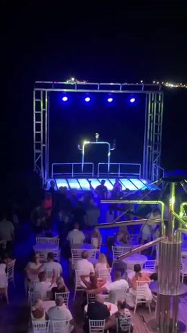 A stage with a DJ booth is illuminated with blue and purple lights. A DJ is visible behind the booth. Below the stage, a group of people are seated at tables and some are standing, watching the performance. The scene appears to be outdoors at night, with a dark background suggesting it might be a boat or a venue near water. The overall atmosphere is that of a party or event.
