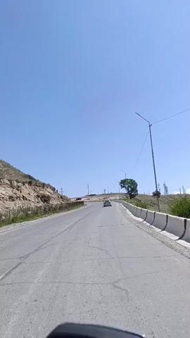 The video shows a view from a vehicle driving on a road. The road appears to be in a rural or mountainous area, with hills and mountains visible in the background. The sky is clear and blue. There are power lines and streetlights along the side of the road. A single car is visible driving in the distance. A concrete barrier lines the right side of the road. The vehicle appears to be moving forward.
