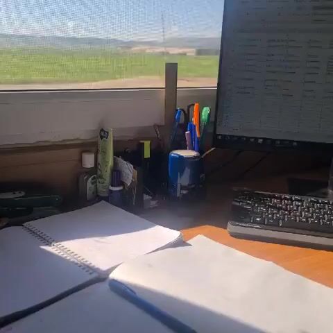 The video shows a desk with a notebook, calculator, stapler, and various writing utensils. Behind the desk is a window with a view of a green field under a blue sky. There is a walkie-talkie on the desk. The scene appears to be an indoor workspace with an outdoor view.