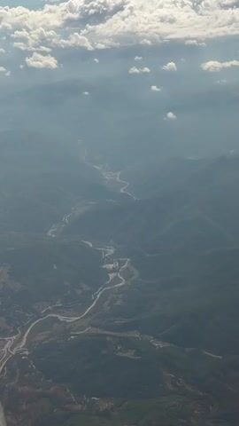 This is an aerial view of a mountainous region with a river winding through it. The sky is partly cloudy with patches of sunlight breaking through. The mountains are covered in green vegetation and appear to be covered in a light haze. There is a road or highway visible alongside the river in some parts of the video. The camera appears to be moving, providing a panning view of the landscape. There are no people or man-made structures clearly visible other than the road. The overall impression is one of natural beauty and vastness.