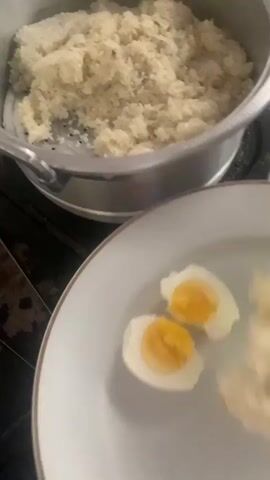 A close-up shot shows a person's hands using a light green utensil to scoop cooked rice from a large metal pot onto a white plate. The plate already contains two halves of a hard-boiled egg, displaying their white and yellow centers. The rice in the pot appears to be white, with a slightly clumpy texture. The lighting is bright, suggesting an indoor setting, possibly a kitchen. In the background, other kitchen items, including another pot and some food items, are partially visible. The video focuses on the action of serving the rice, highlighting the preparation of a simple meal.