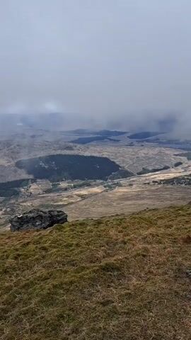 The video shows a landscape from a high vantage point with two people hiking on a grassy hill. The landscape below is expansive, with patches of green and brown terrain and visible bodies of water in the distance. The sky is overcast with low-hanging clouds obscuring parts of the view. The two individuals are wearing hiking gear, including backpacks and waterproof jackets, and are walking away from the camera along the ridge of the hill. The overall atmosphere is one of adventure and remote natural beauty, despite the gloomy weather.