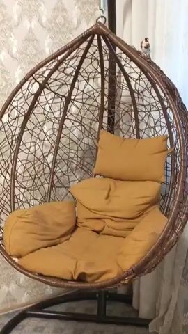 The video shows a hanging egg chair with tan cushions.  A small bird with a red face, brown and white body is perched on the edge of the chair, close to the top. The bird is looking up and towards the left.  A white curtain is visible in the background.  The camera zooms in on the bird.