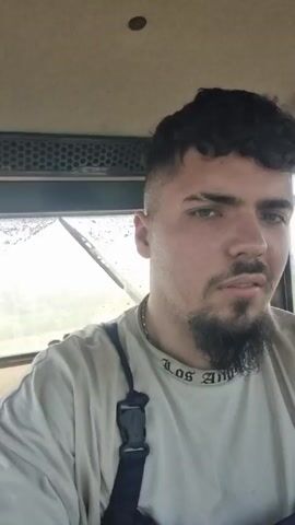 The video shows a man inside the cab of a tractor, which is pulling a red agricultural implement in a field. The man has dark hair, a beard, and is wearing a light-colored t-shirt with the words "Los Angeles" on the collar, along with dark-colored overalls. The view through the front window of the tractor reveals dusty conditions as the tractor moves through the field. The agricultural implement consists of a series of wheels, possibly discs, used for tilling or preparing the soil. The tractor cab is partially visible, with a black vent or grill near the ceiling.