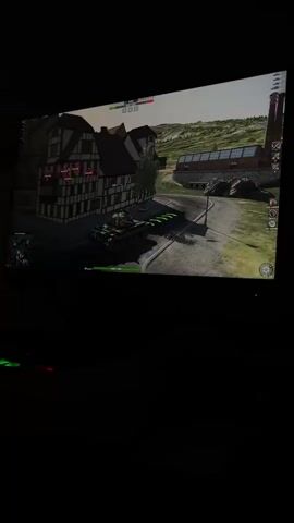 A person plays a video game on a large screen. The game appears to be a tank combat simulation, with a tank moving through a village environment. The screen displays game-related graphics, including health bars and aiming reticles. The room is dark, with only the light from the screen illuminating the scene.
