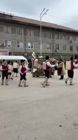 A group of people in traditional Bulgarian clothing are performing a ritualistic dance in a public square. They are wearing black hats, white shirts, red vests, and dark pants, with bells attached to their waists. They are walking in unison, striking the bells in time with the music. In the center of the group, there is a large figure covered in straw or fabric. The dance is taking place in front of a large, multi-story building, with a crowd of people watching. A tent and some balloons in the colors of the Bulgarian flag are visible in the background. The sky is overcast.