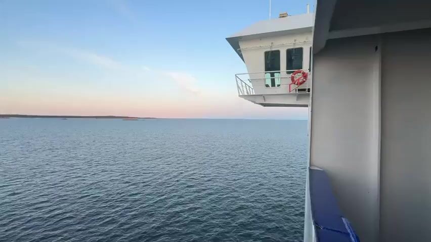 The video shows a view from a ship sailing on the sea. On the right side of the frame, part of the ship's superstructure is visible, including a white bridge with windows and a railing. The sea is calm with small waves, and the sky is a gradient of blue and pink, suggesting either sunrise or sunset. In the distance, the horizon shows a faint outline of land or islands. The overall scene is serene and depicts a journey at sea.