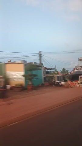 The video is a shaky, motion-blurred recording of a roadside scene, likely from a vehicle in motion. The sky is visible in the upper portion of the frames, appearing to be either early morning or late evening with a soft blue hue. The ground in front of the buildings is a reddish-brown dirt or gravel. The buildings appear to be a mix of commercial structures and residences, with some showing signs of habitation or small businesses. In the foreground, a reddish-brown road surface is visible.

In the first frame, a building with a light-colored facade and a turquoise gate is on the left. Several motorcycles are parked on the side of the road. Further down the road, there are more buildings and what appears to be a market or roadside stalls with people and goods displayed. Power lines are strung across the top of the scene.

The second frame shows a white building with red trim, possibly a restaurant or shop, with an open-air seating area. A water tower is visible in the background.

The third frame shows a white building with dark windows, adjacent to what looks like a more industrial or utilitarian structure with a large, dark opening. There is a red and white striped post or marker on the right side of the frame.

The fourth frame shows a dense green tree with a large structure behind it, possibly a warehouse or large building with a corrugated metal roof. Piles of what appear to be trash or debris are visible on the ground in front of this structure.

The final frame shows a blue metal gate on a white building, with some greenery and a pile of orange bricks or blocks to the right.
