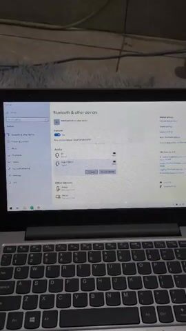The video displays a laptop screen showing Windows settings for Bluetooth devices. A small red fan shaped like an Angry Bird is visible in the background. The fan is plugged in and appears to be on, with its blades spinning. The environment is dimly lit and appears to be a room with a rug on the floor.