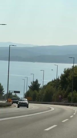 The video shows a view from a car traveling on a highway overlooking a body of water and distant hills. Several cars are visible on the road, including a dark-colored SUV and a lighter-colored sedan. Streetlights line the side of the highway, and there are trees and vegetation on the slopes. The sky is clear and blue, with some faint clouds. In the distance, a person can be seen parasailing over the water. The scene appears to be a sunny day.