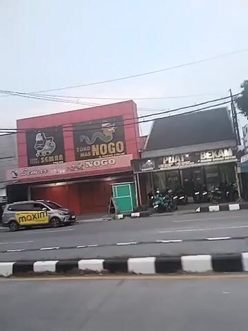 The video shows a street scene. In the initial shot, a building with the word "NOGO" on it is visible, and traffic moves in the foreground. The camera pans to the left, showing more storefronts and parked motorcycles. It continues to pan left, ultimately revealing a large building with the word "matahari" visible. The day appears to be overcast. There are a few people present, but the focus is on the buildings and traffic.