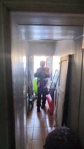 A person in a patterned jacket walks toward the camera through a narrow hallway. The walls of the hallway are lined with reflective panels. To the right are cleaning supplies and a leaning door. A sign with the word "EXIT" in Russian is visible above the exit door at the end of the hallway.
