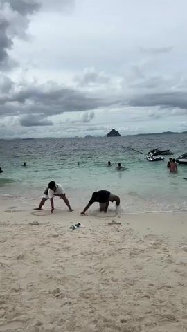 The video shows a beach scene with two young men in the foreground interacting with the sand and water. The man on the left is wearing white shorts and a white t-shirt and appears to be drawing in the sand with his finger. The man on the right is wearing dark shorts and is partially submerged in the water, with his head down as if digging or playing in the sand at the water's edge. A pair of flip-flops lies on the sand between them. In the background, several other people are in the water, some swimming and others wading. Further out, a jet ski is visible, and a small, hilly island is in the distance. The sky is overcast with dark, dramatic clouds.