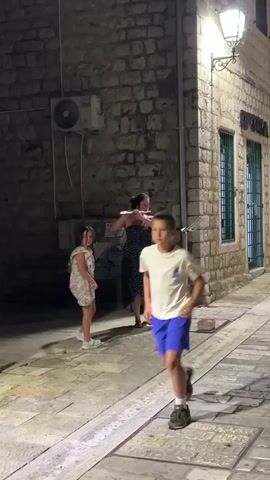 A young boy in a white t-shirt and blue shorts runs down a cobblestone alleyway at night, his motion blurred by the camera. Behind him, a woman in a black polka-dot strapless dress stands playing a flute. She is positioned next to a music stand. To the left of the woman, a young girl in a white dress with ruffles stands looking forward. The alley is lit by a street lamp and ambient light reflecting off the stone walls and buildings. The scene appears to be outdoors in a European-style town.