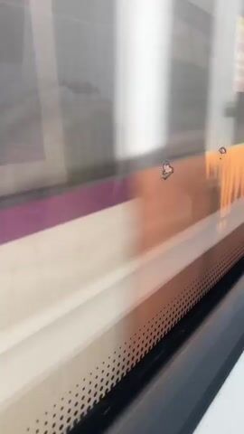 The video shows a close-up view through a window, possibly of a train or bus. The window has a purple stripe running horizontally across it. The exterior is blurred, indicating movement, and features a light-colored surface with a darker, patterned strip at the bottom. There are some small imperfections or marks on the surface of the window. The lighting is bright, suggesting daytime.