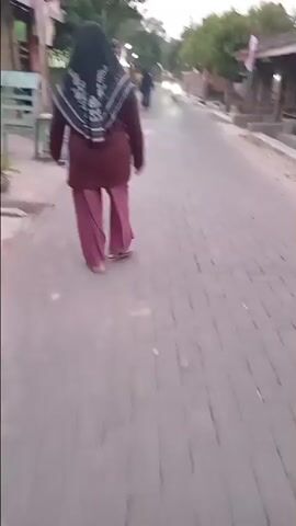 The video begins with a view from behind a person wearing a maroon top, pink pants, and a black and white hijab walking away from the camera. The person is walking on a paved path made of bricks. To the left of the path are some plants and what appears to be a small building or structure. To the right of the path, the path continues and trees can be seen. As the video progresses, the person continues walking down the path. A motorcycle with a rider passes the person from behind, heading in the same direction. Another motorcycle can be seen in the distance further down the path. The environment appears to be a residential street or alleyway, possibly in a tropical or sub-tropical climate, given the greenery. The lighting suggests it is either early morning or late afternoon. The video continues to show the person walking away from the camera, with the environment remaining consistent.